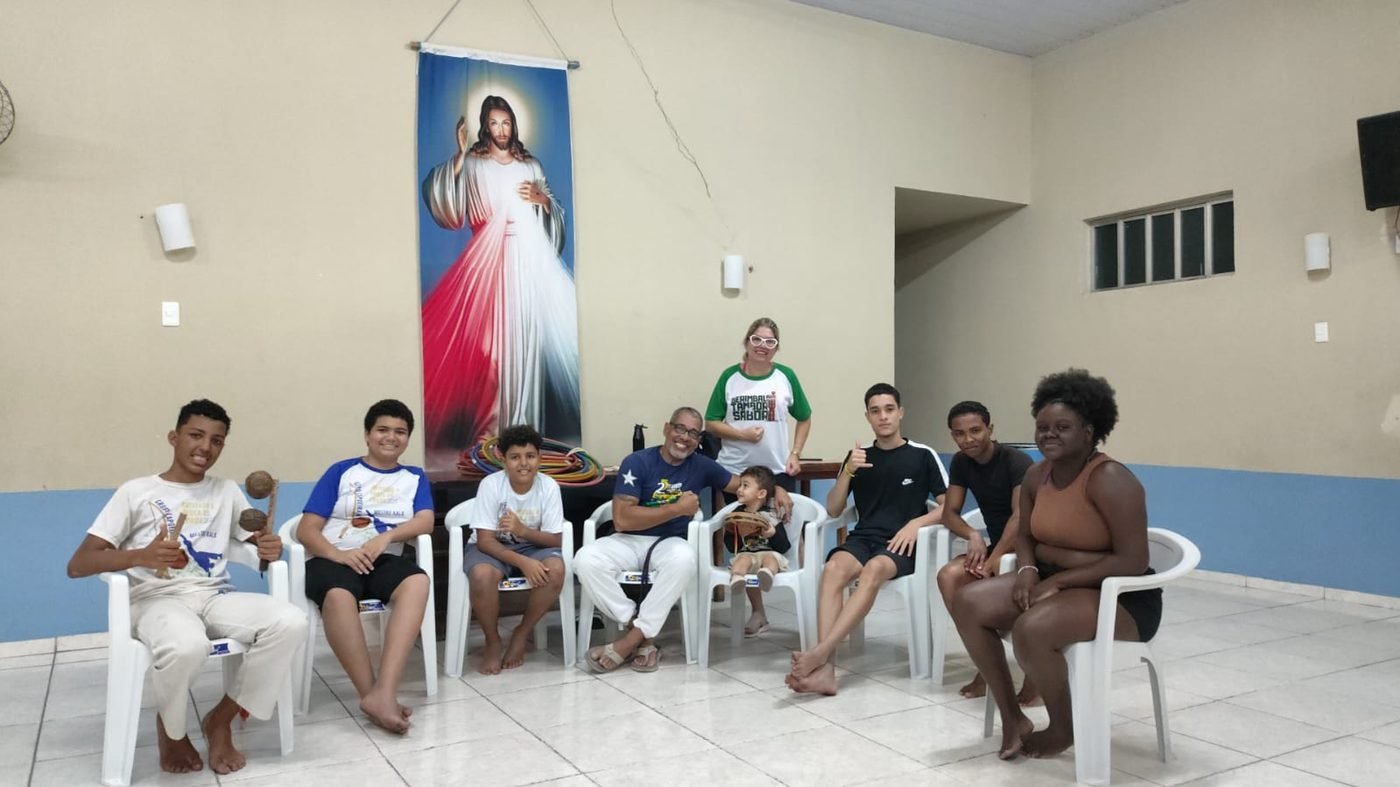 Crianças e voluntários sentados em roda com instrumentos de capoeira durante oficina cultural do instituto